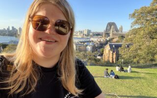 Nainen aurinkolasit päässä hymyilee ja taustalla on Sydneyn satamasilta kuvattuna Sydneyn Observatorio Hilliltä